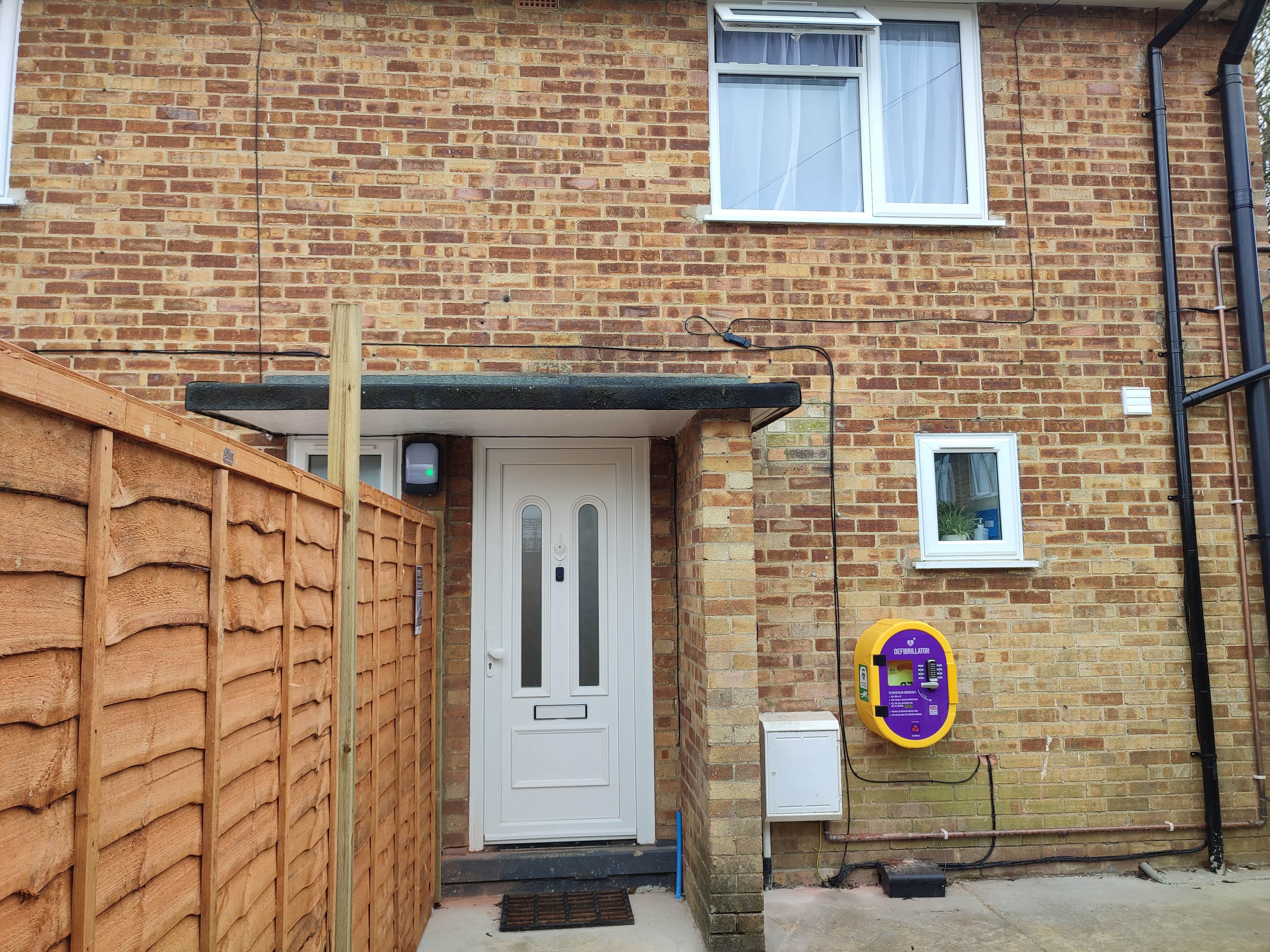 A photo of the outside of a house. It is made of red bricks and it two stories tall. There is a small porch covering the white front door. There are two windows visible on the front of the house. One one the top floor and one one the ground floor. Underneath the ground floor window is a yellow and purple defibrillator box. 
