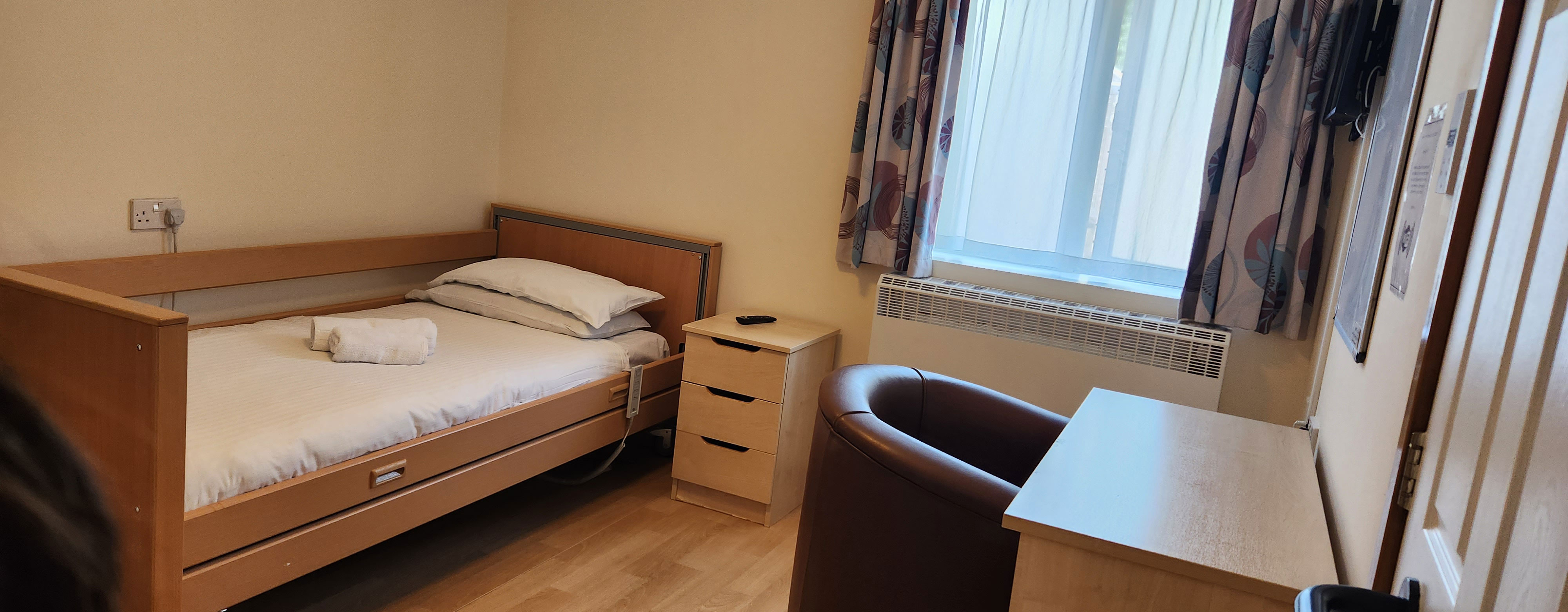 A bedroom. there is a single bed ion the left hand side of the room. A window is on the back wall and there is a desk on the right with a leather arm chair tucked underneath it