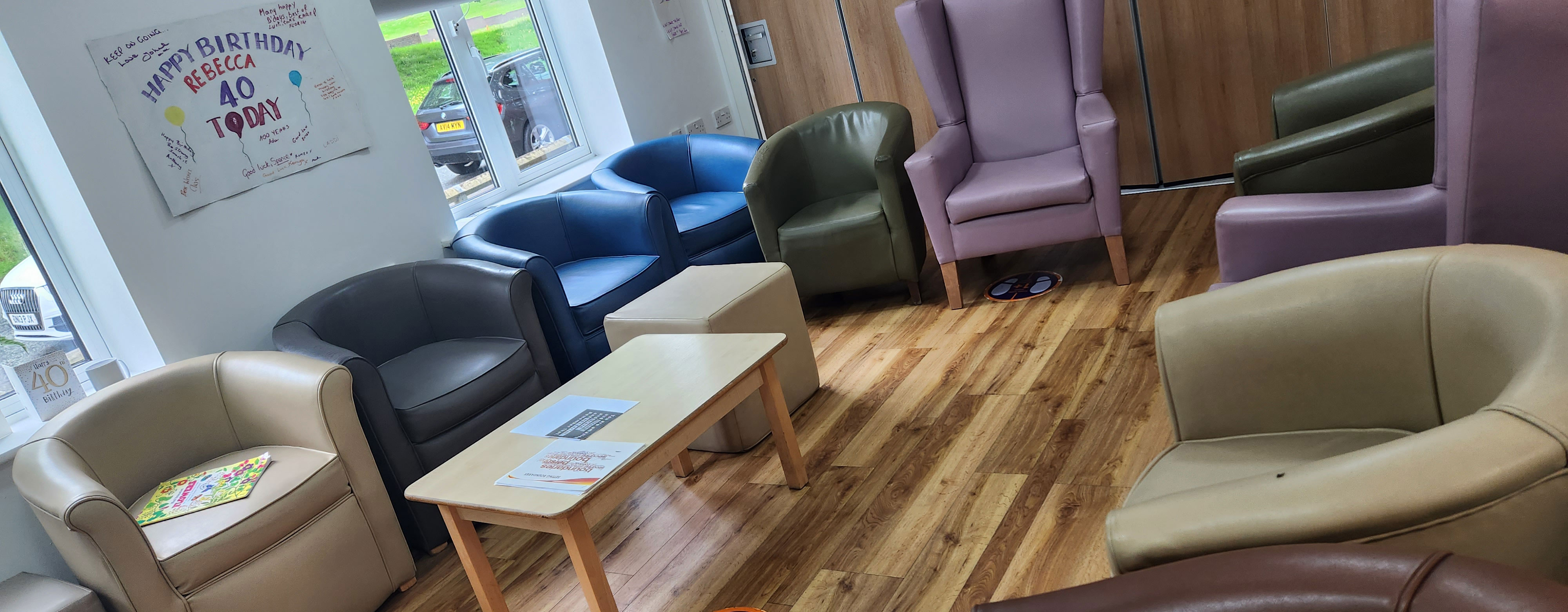 A communal area. there is wooden floors and two windows letting a lot of light in There are a selection of different arm chairs dotted around the edge of the room.