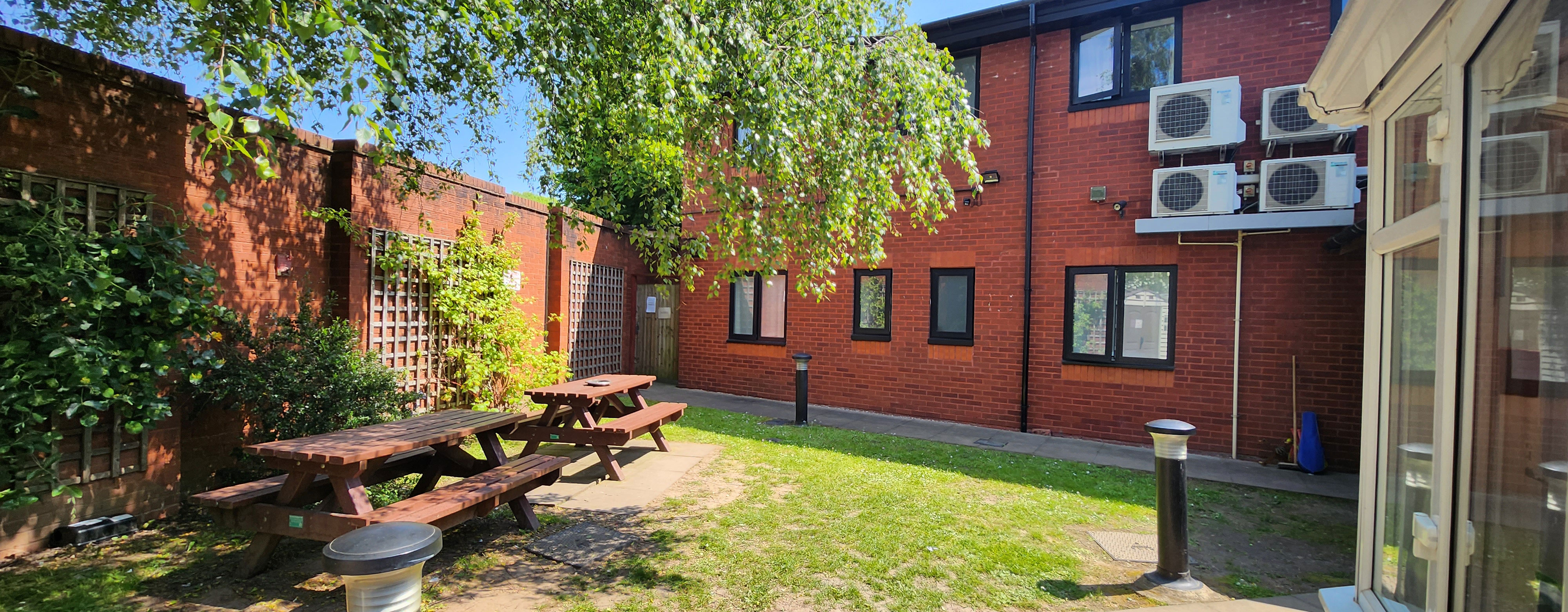 A garden. The Park House building is in the back of the shot with red brick walls. there is a brick wall along the left hand with trellis along the wall. There are two wooden picnic benches underneath a tree.