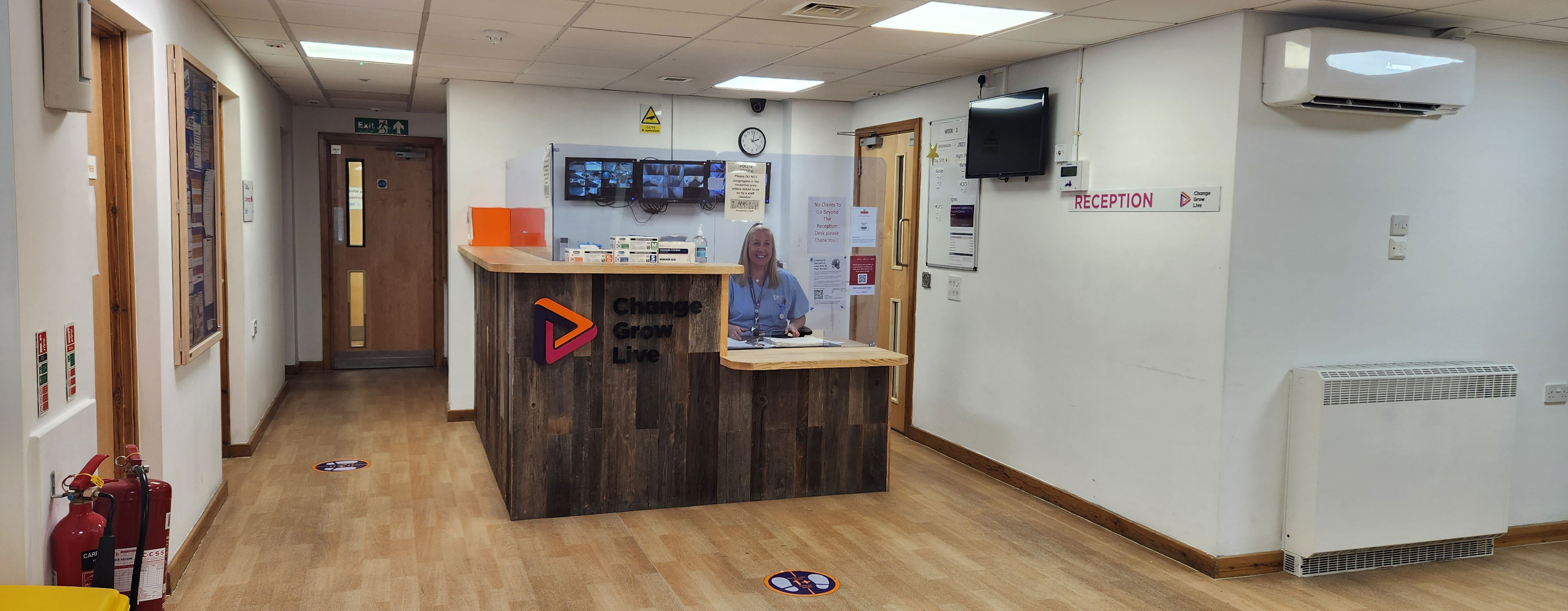 The park house reception area. There is a desk in the middle of the room with perspex glass around it. There is a friendly person sat behind the desk smiling at the camera.