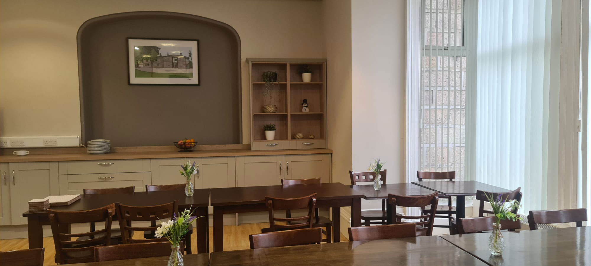 A photo of a dining room. There are 2 rows of dark brown tables with chairs tucked underneath them. There are cupboards and counterspace behind the tables.