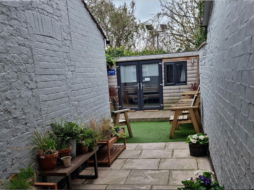 A Photo of a courtyard. There's a patio in the foreground, white walls either side, some grass and a garden office at the far end
