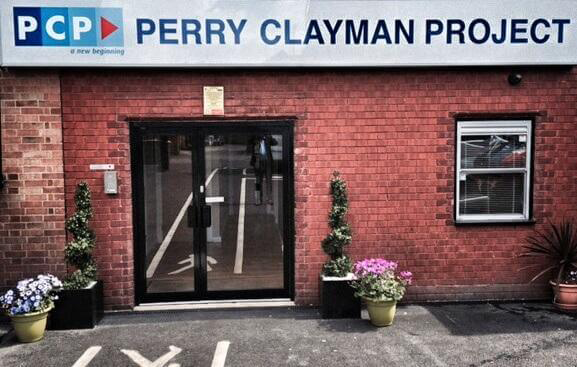 A red brick wall with a double glass door. A Perry Clayman Project sign hang above