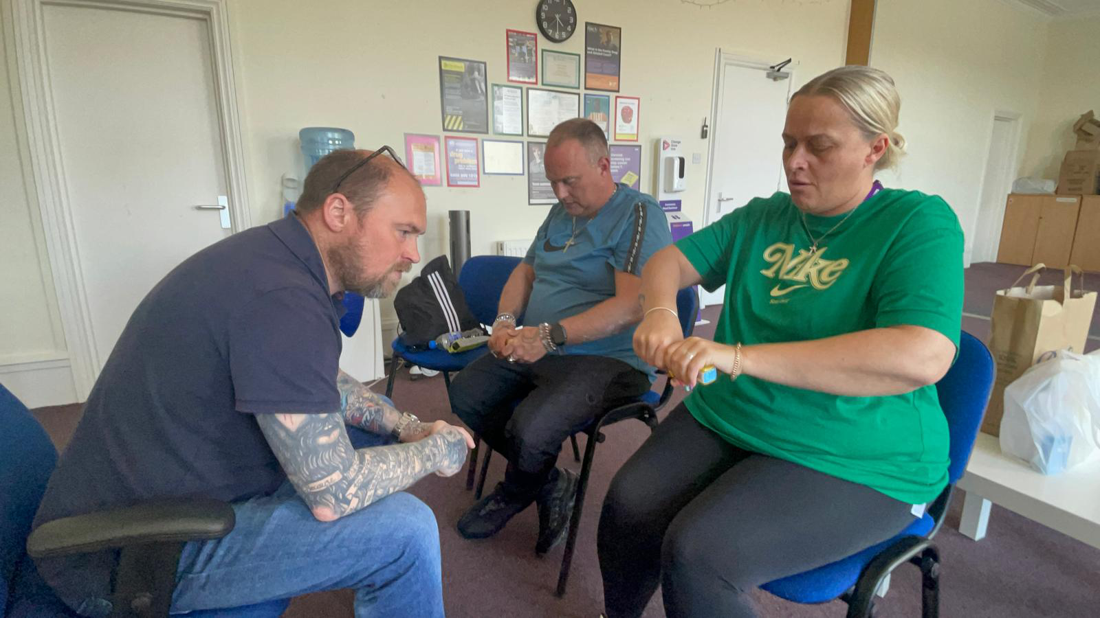 Three people sat on chairs, one is facing the other two. They are holding yellow Naloxone boxes