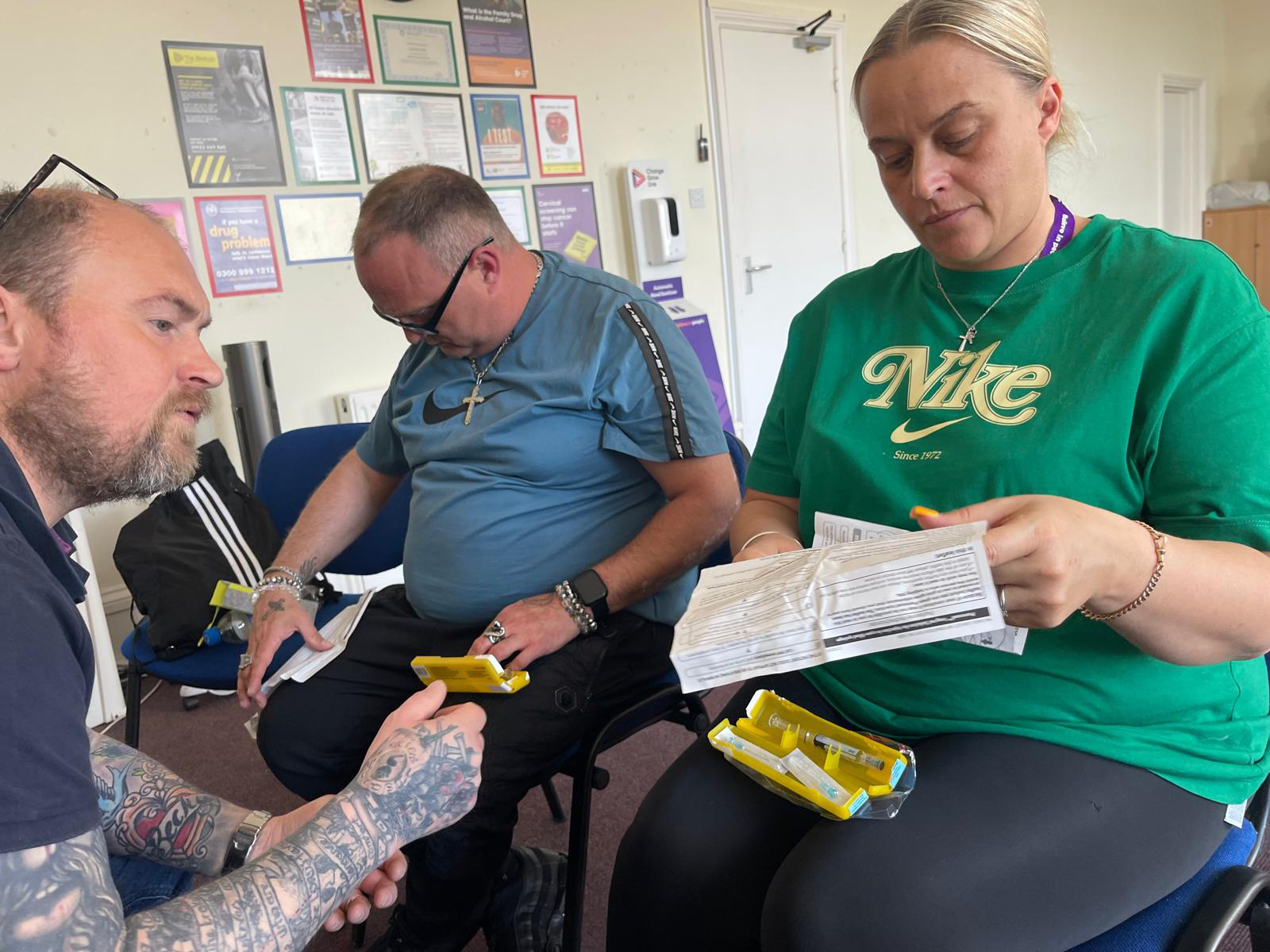 Three people sat on chairs, one is facing the other two. They are holding yellow Naloxone boxes