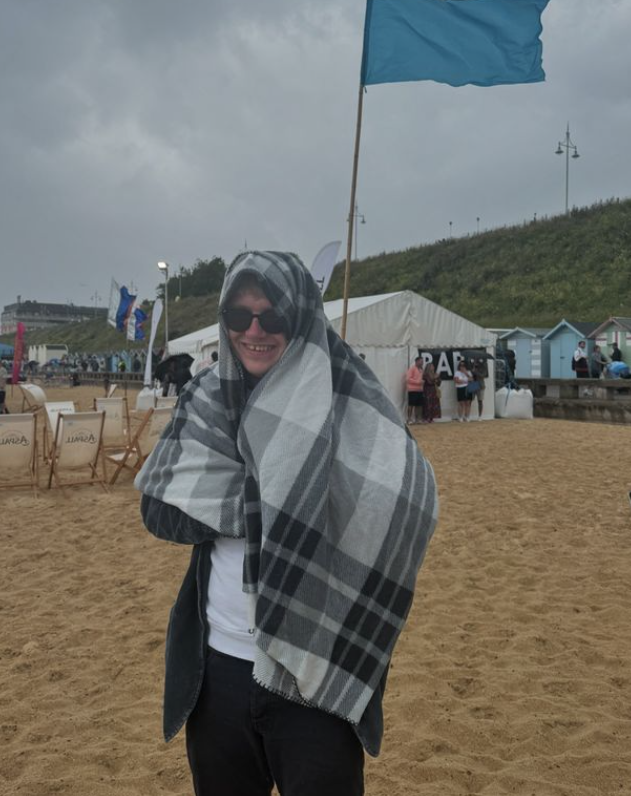 A young man stood on a beach with a blanket wrapped around him