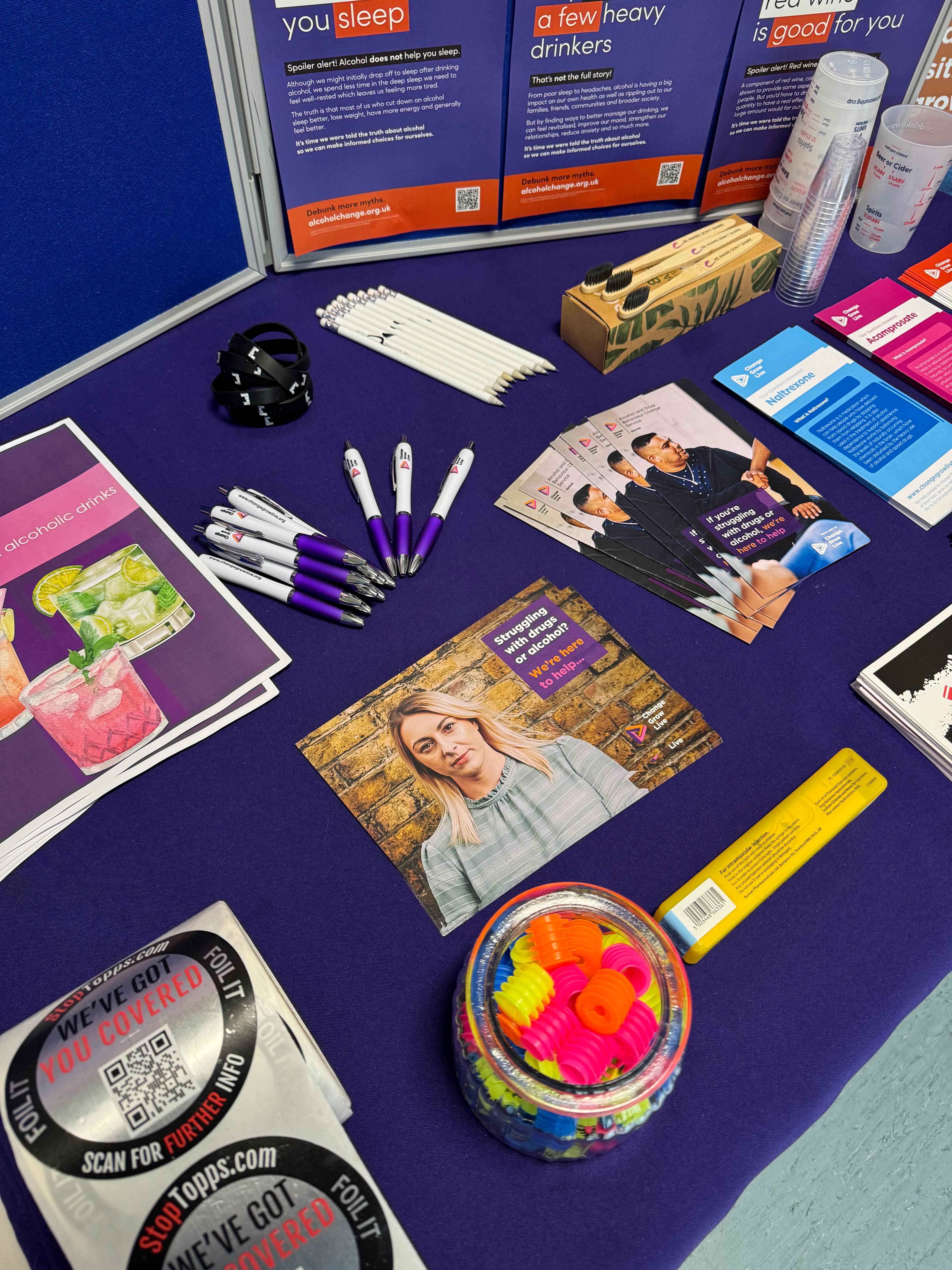 A close up of a table with stickers, leaflets, pens and a naloxone kit on it
