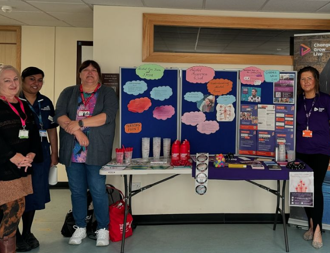 Four people stood either side of a table with a display board with posters and information around alcohol awareness week