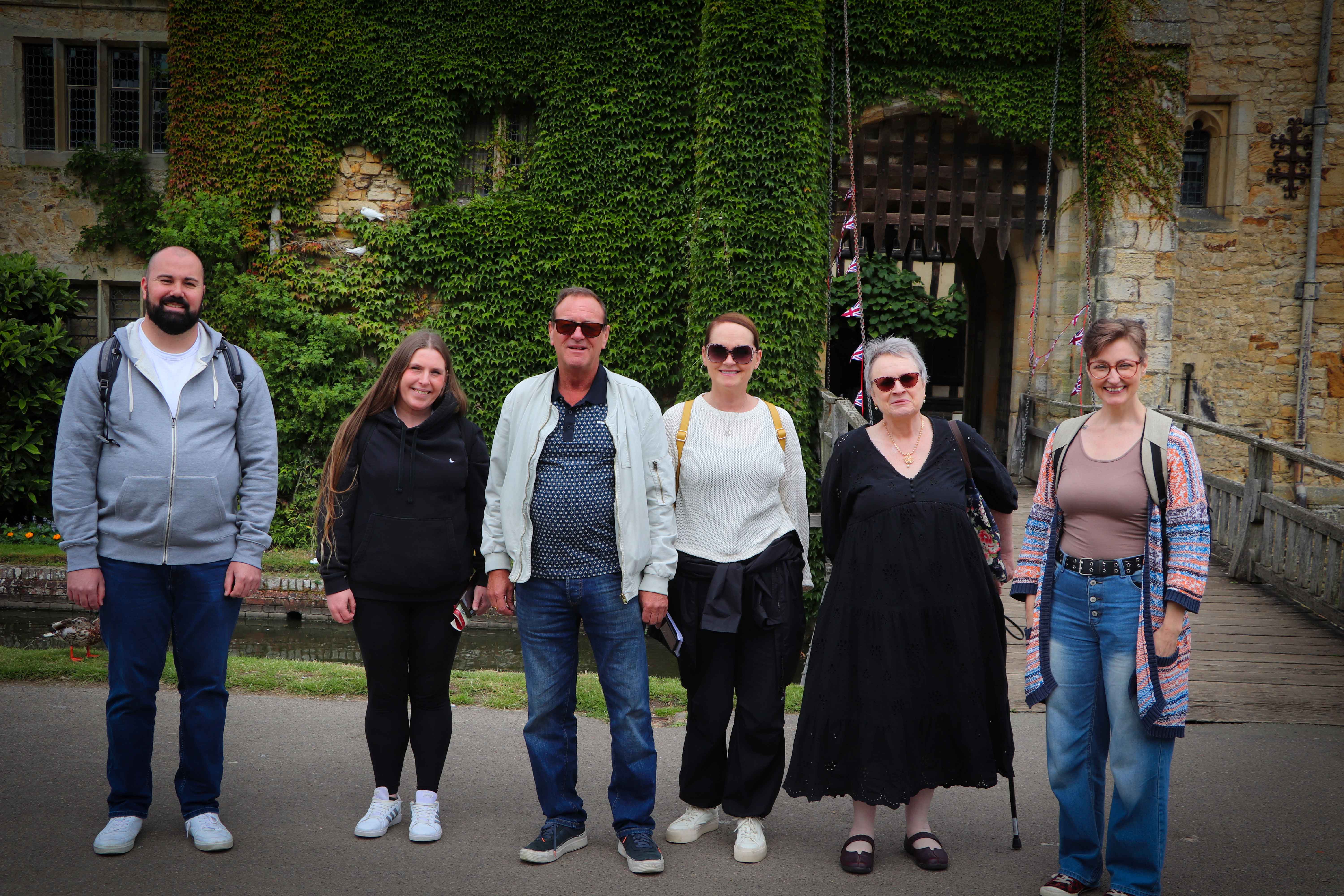 six people stood posing for a photo. Behind them is an old wall with plants trailing up it