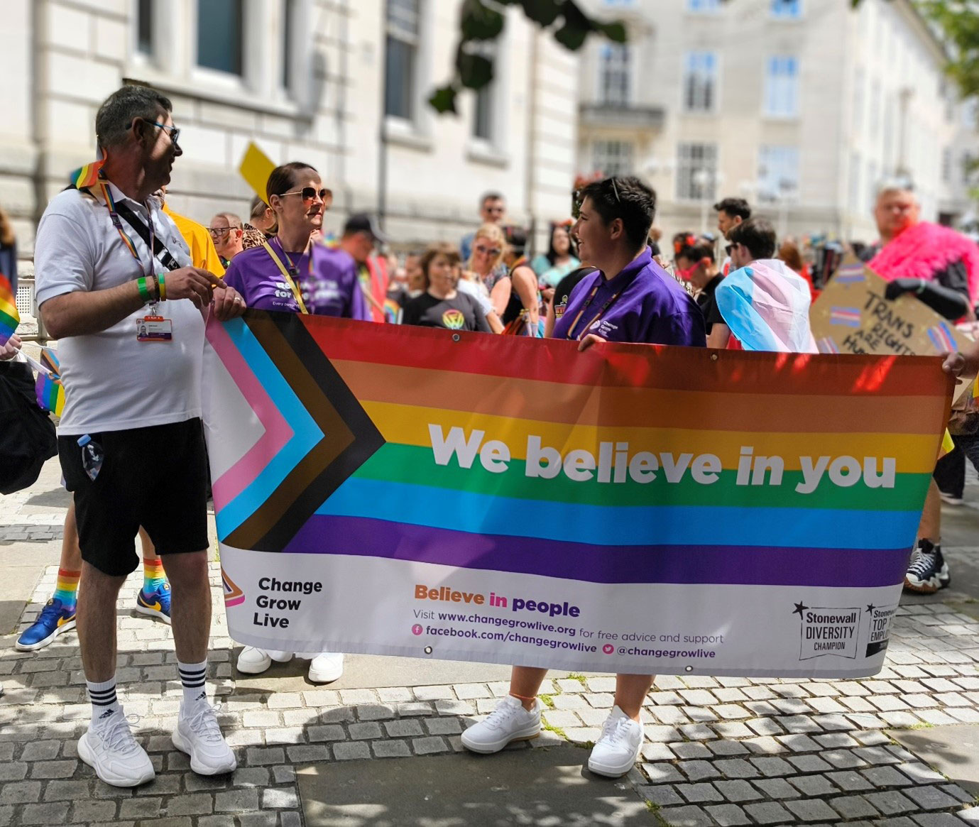 A group of people stood in the shade holding up a pride banner which reads "We believe in you"