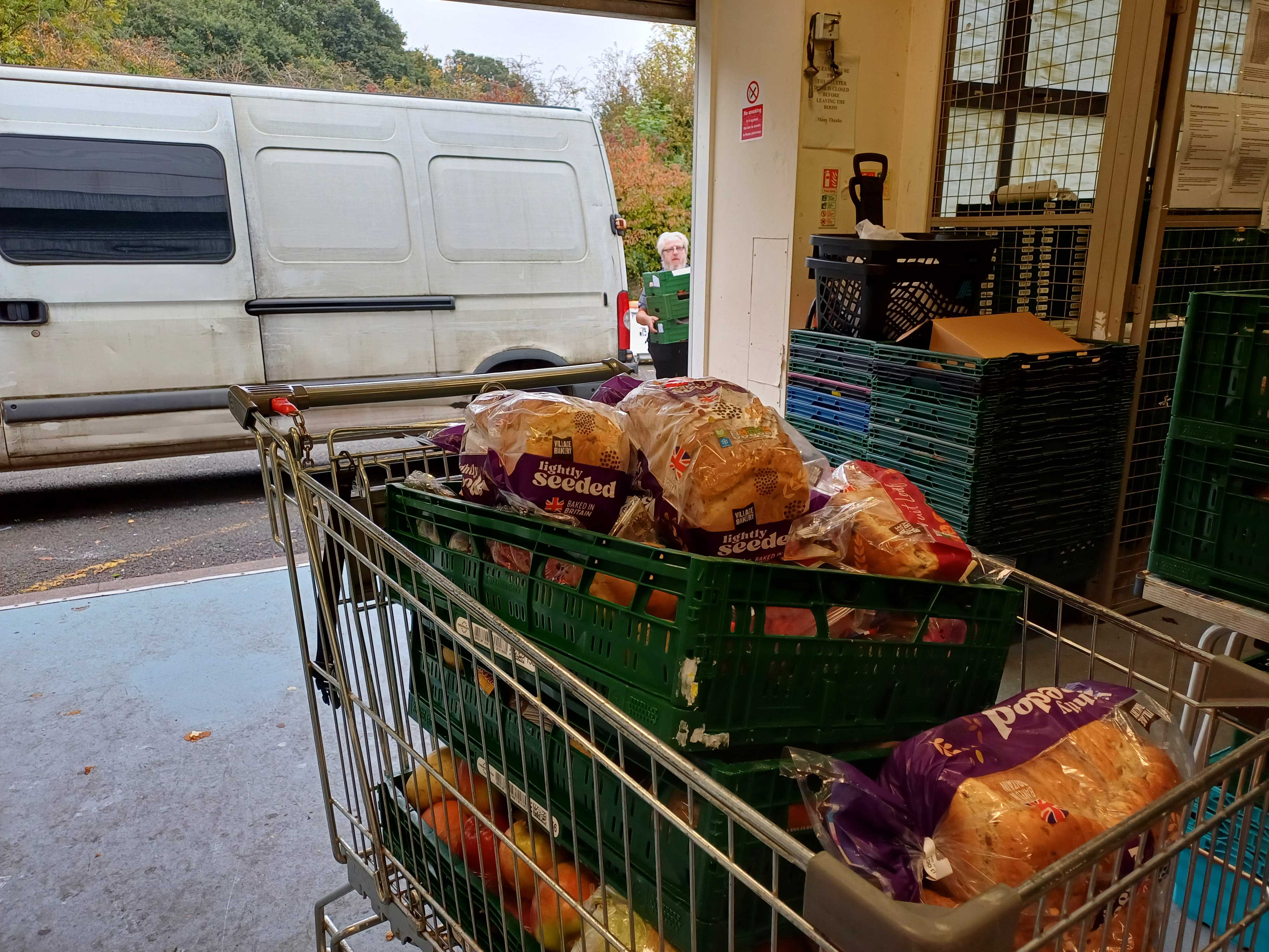 A shopping trolley full of food in the entrance of a warehouse with a white van parked outside