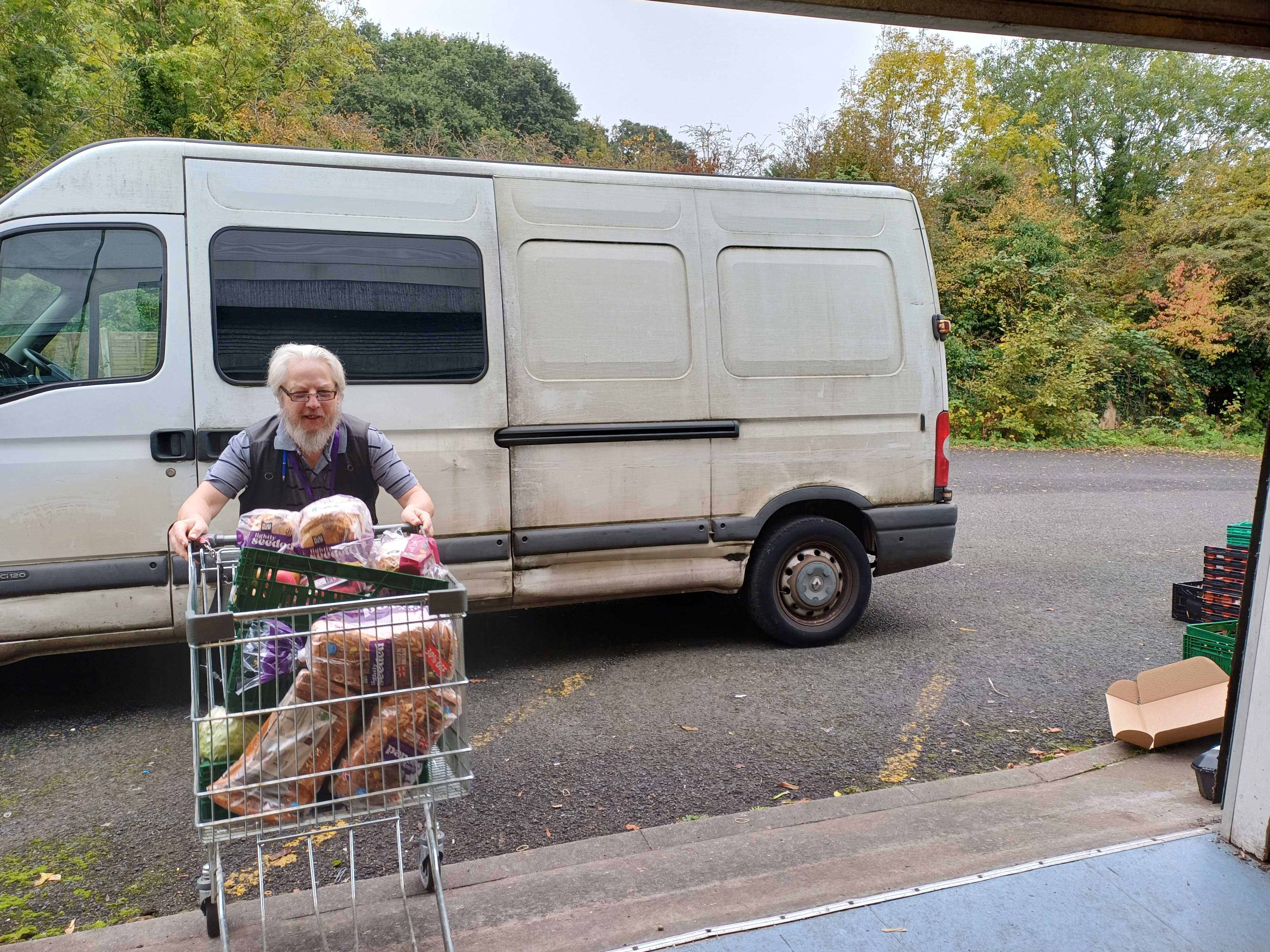 A man pushing a shopping trolley full of food into a warehouse. A white van is parked behind him