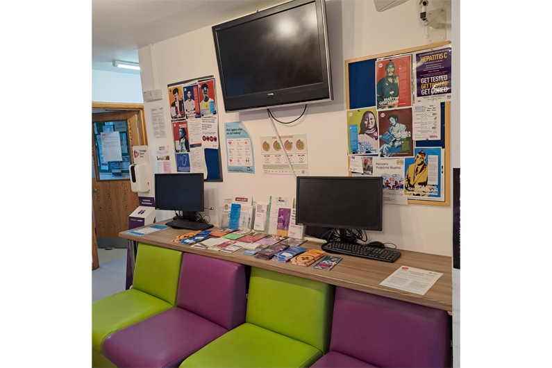 A line of alternating purple and green sofa chairs in front of a desk with two computers and a notice board