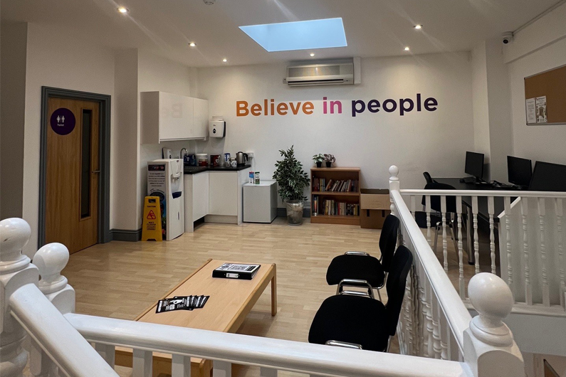 A open waiting area with seats around the edges. A sign with 'believe in people' is handing on the wall.