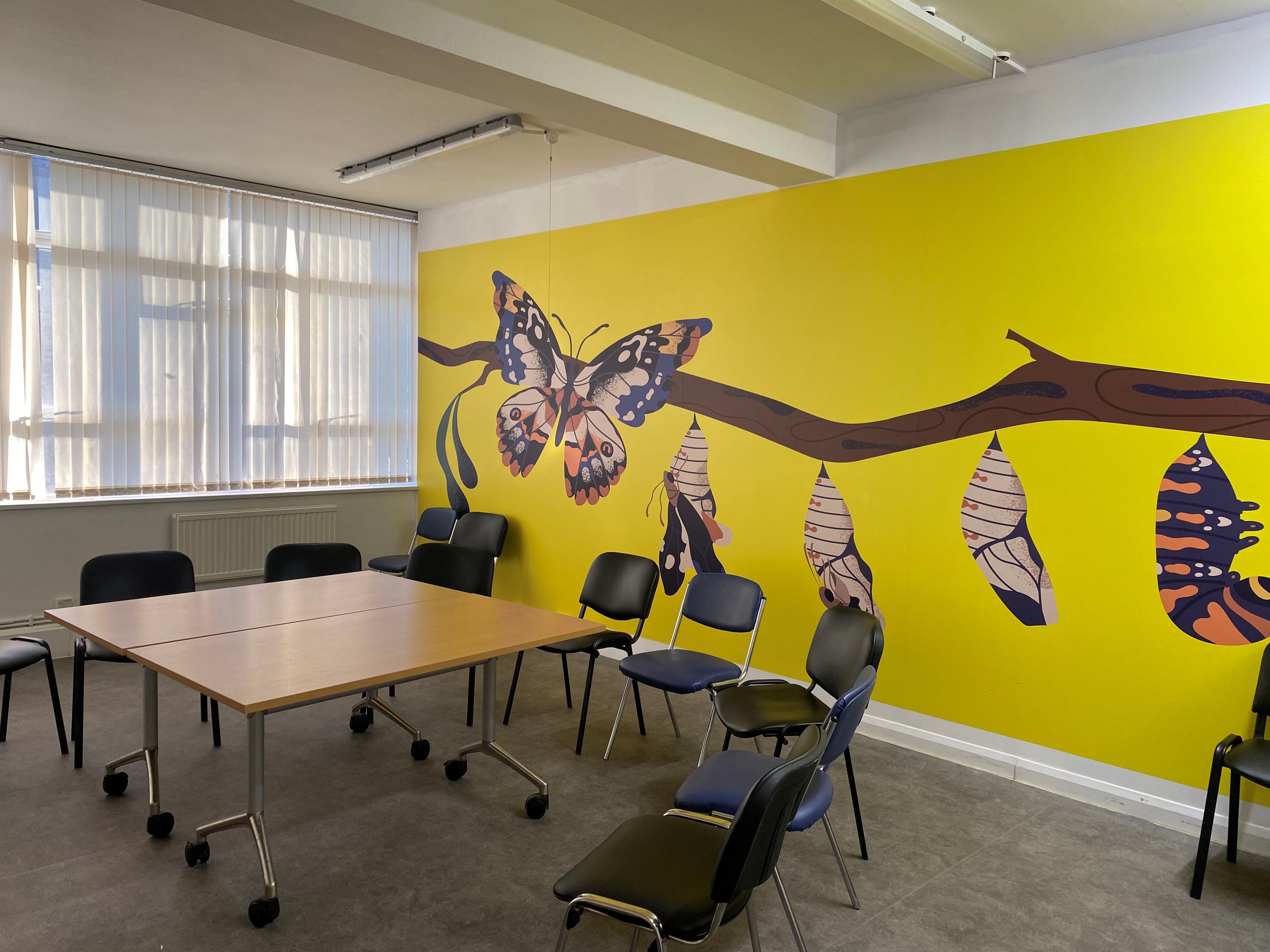 A bright yellow wall with a branch and caterpillar pods transforming into butterflies hanging from it. In the room is a table with chairs around the outside