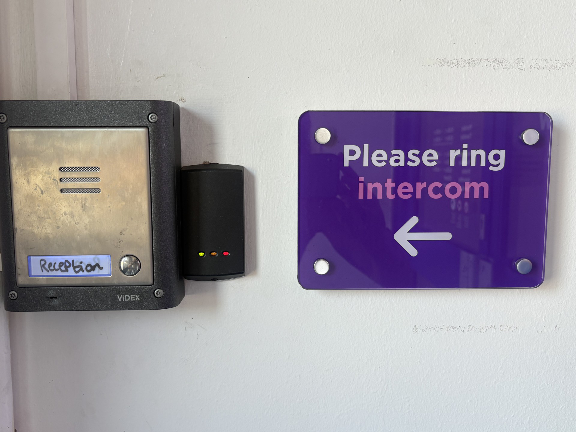 An intercom buzzer mounted to the wall with a sign next to it saying 'Please ring intercom' and an arrow pointing to the left