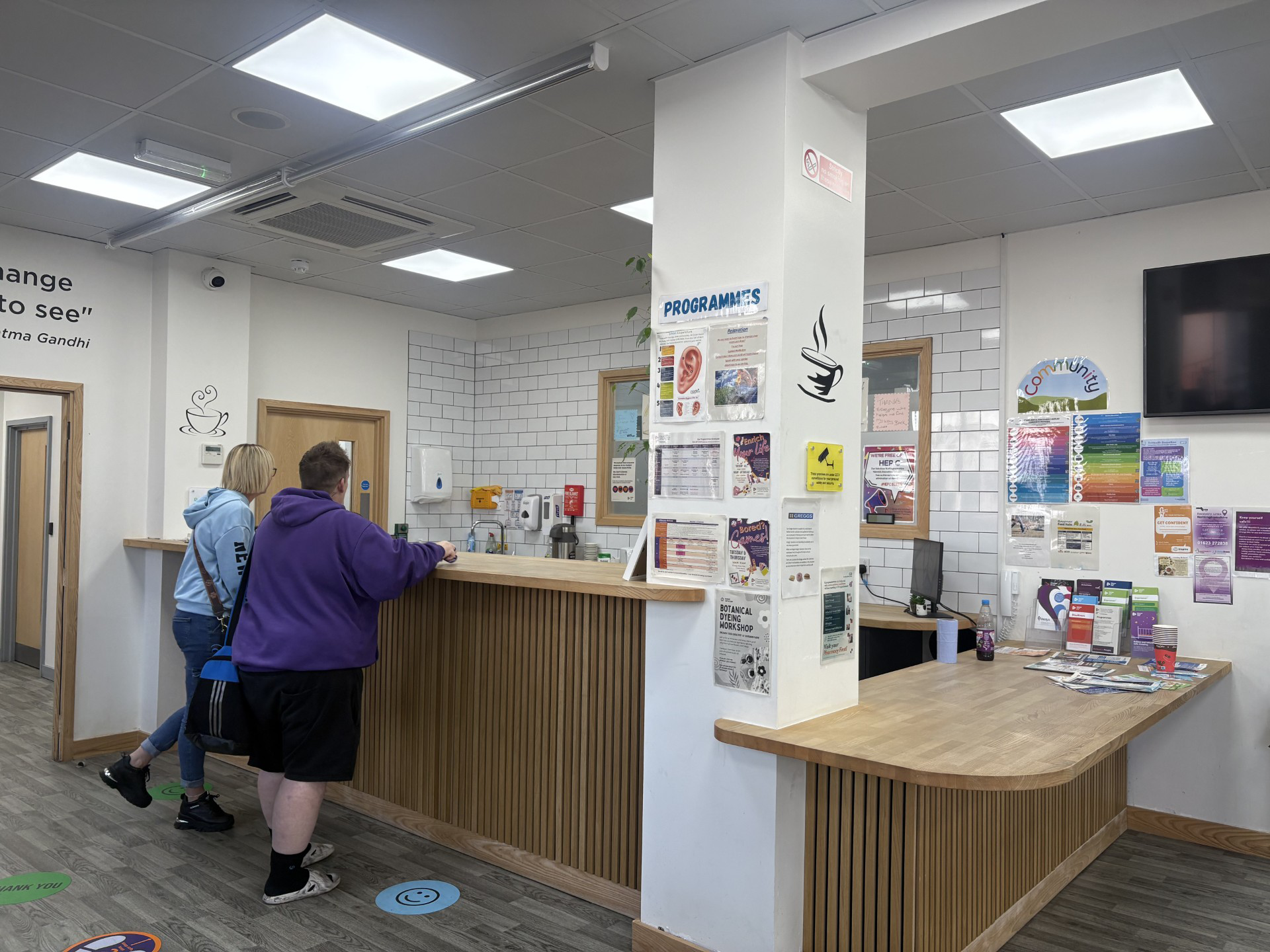 A reception desk with two people leaning on the counter. There are posters and leaflets scattered across the desk