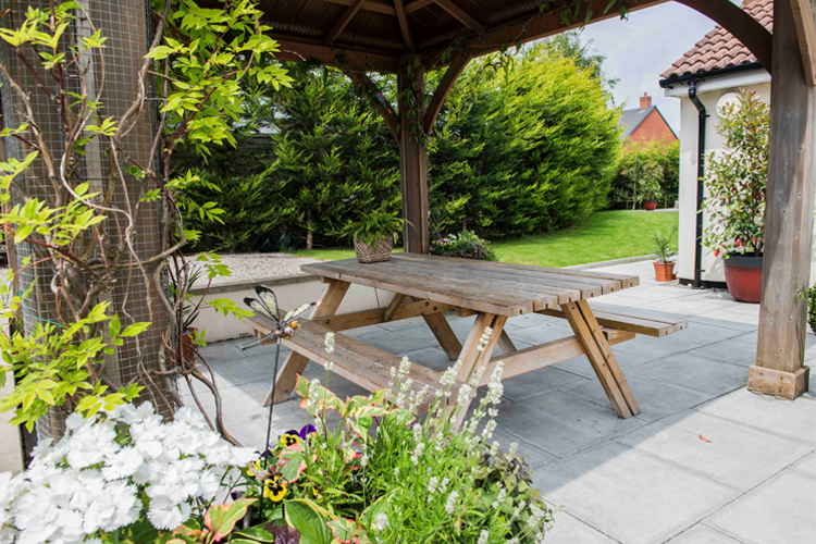 A patio area underneath a wooden gazebo with flowers trailing up it. A wooden picnic table sits in the middle