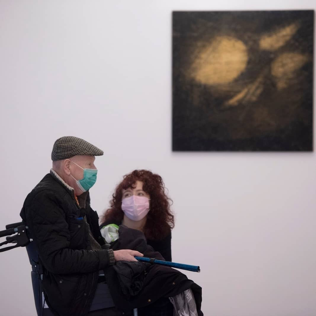 A woman crouching down to speak with an elderly man in a wheel-chair