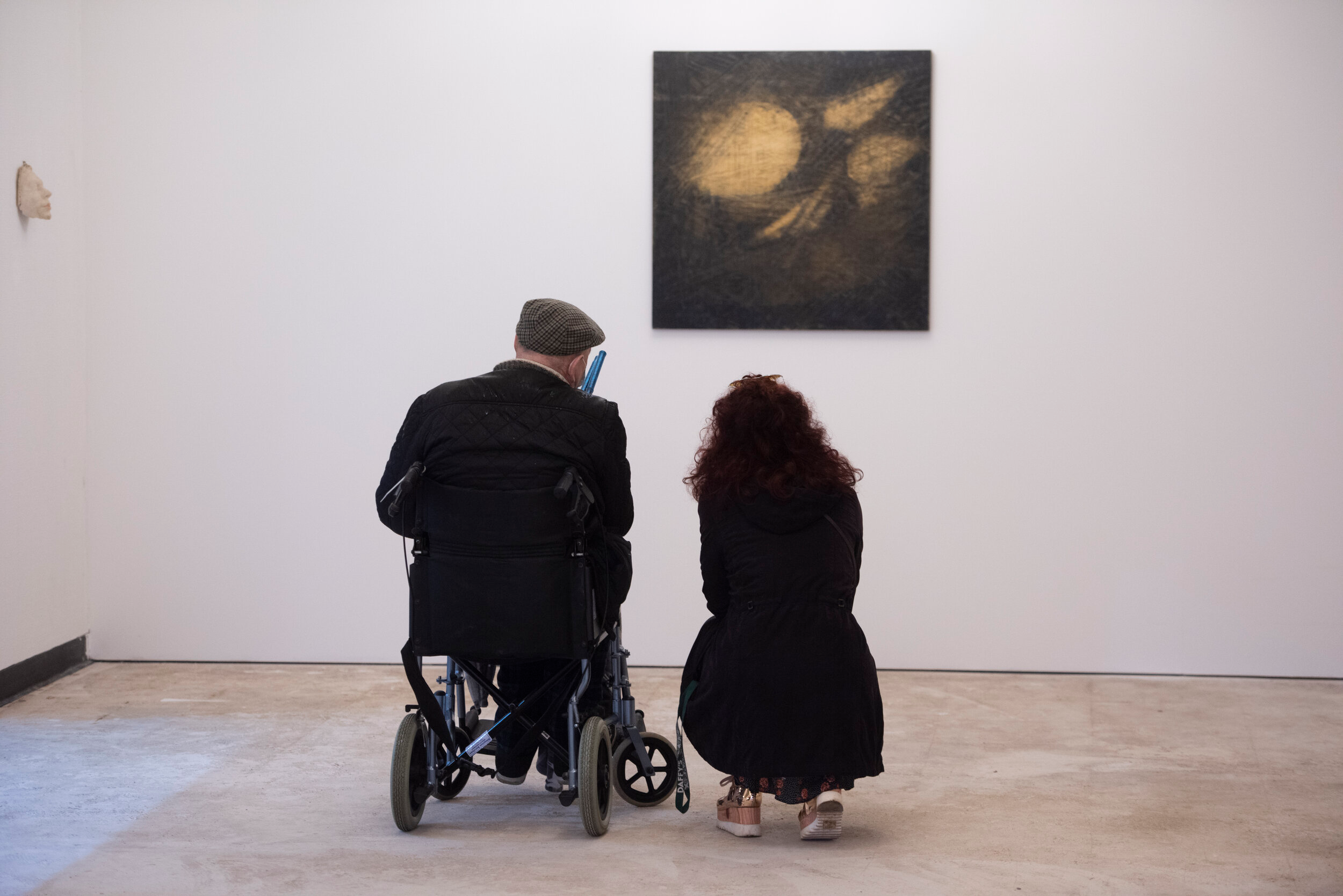 A woman crouched down on the floor next to a man in a wheelchair looking at art on the wall
