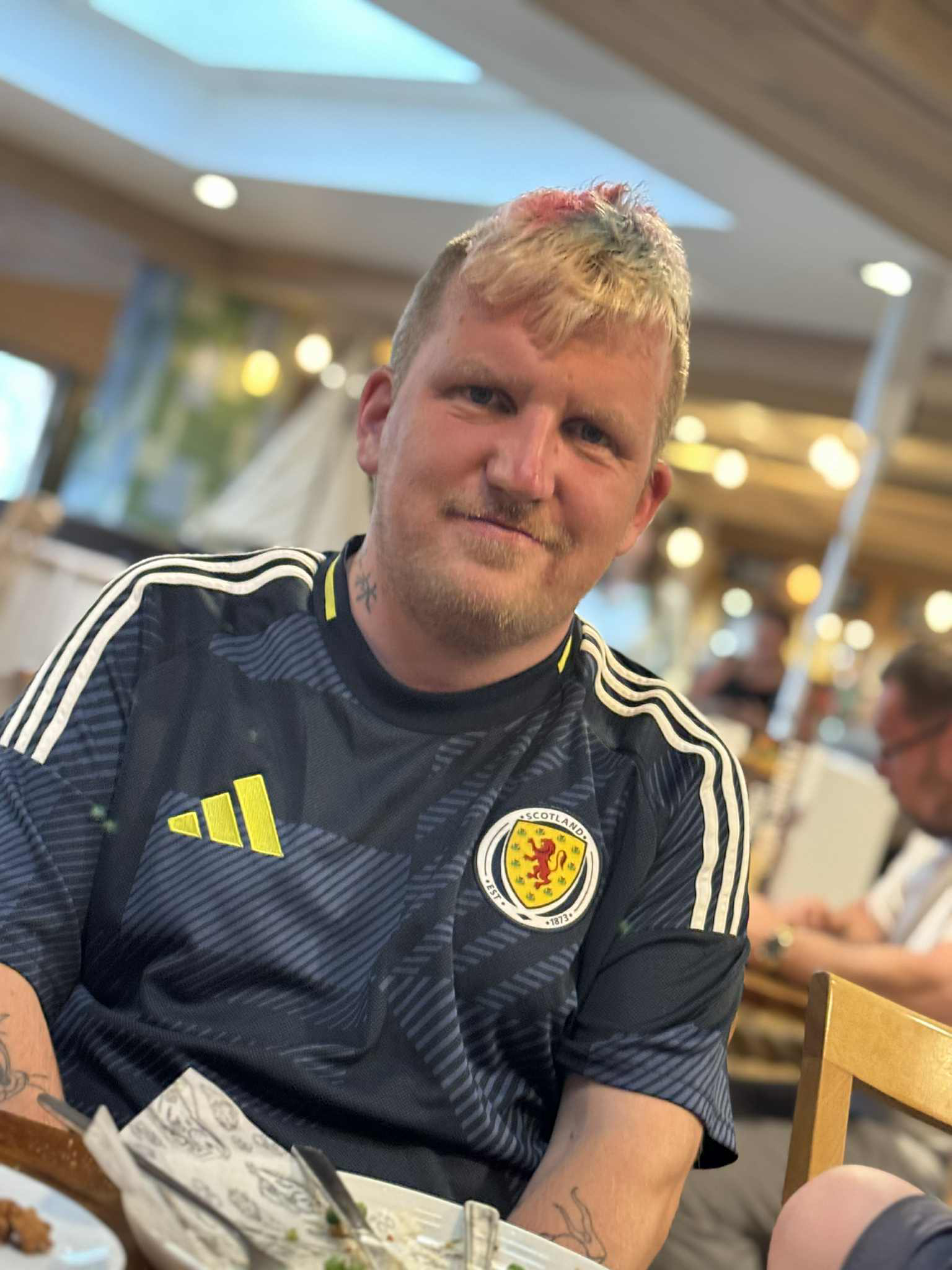 A man with blond hair and a pink streak sat in a restaurant wearing a football shirt