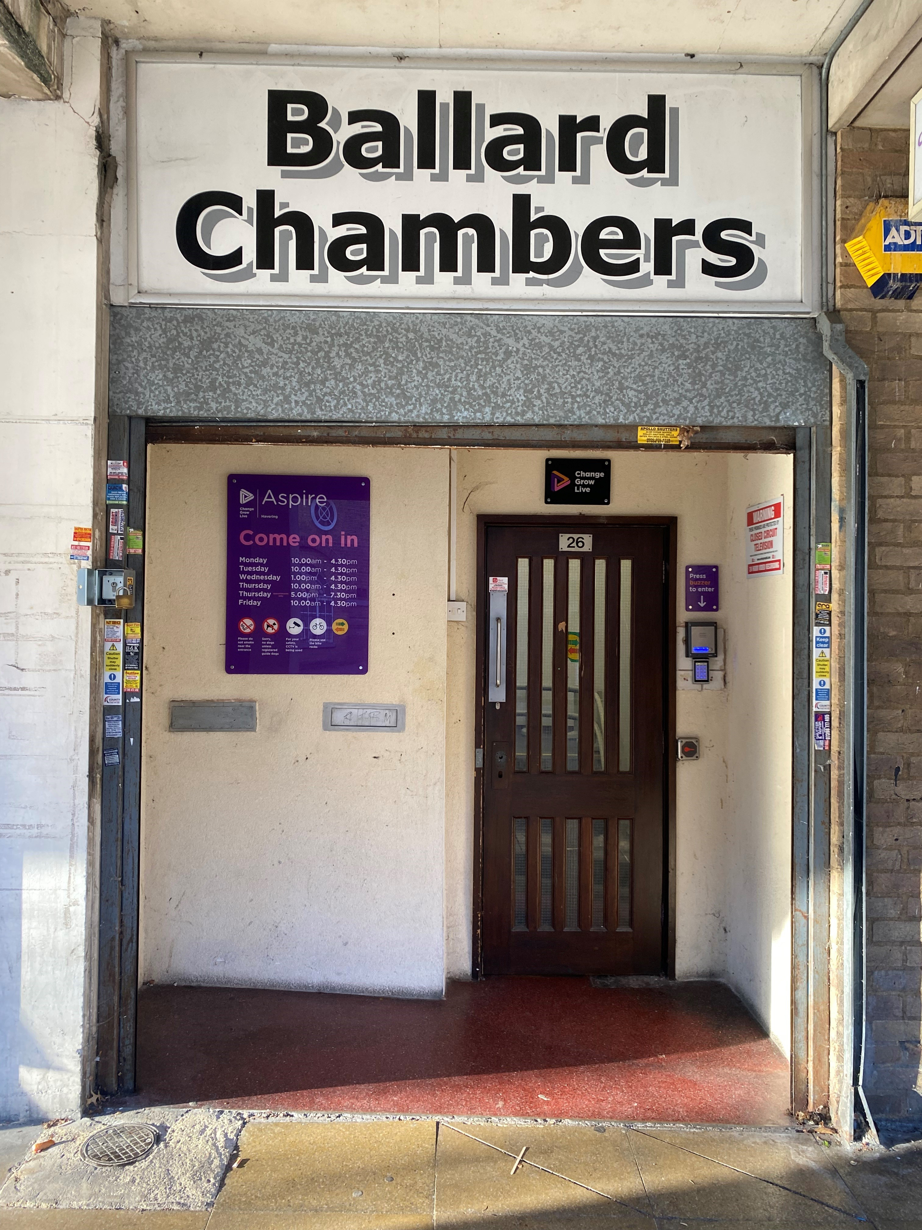A front door inside of a building. Ballard Chambers is written above the entrance and there is a purple sign next to the door with the opening times of the service.