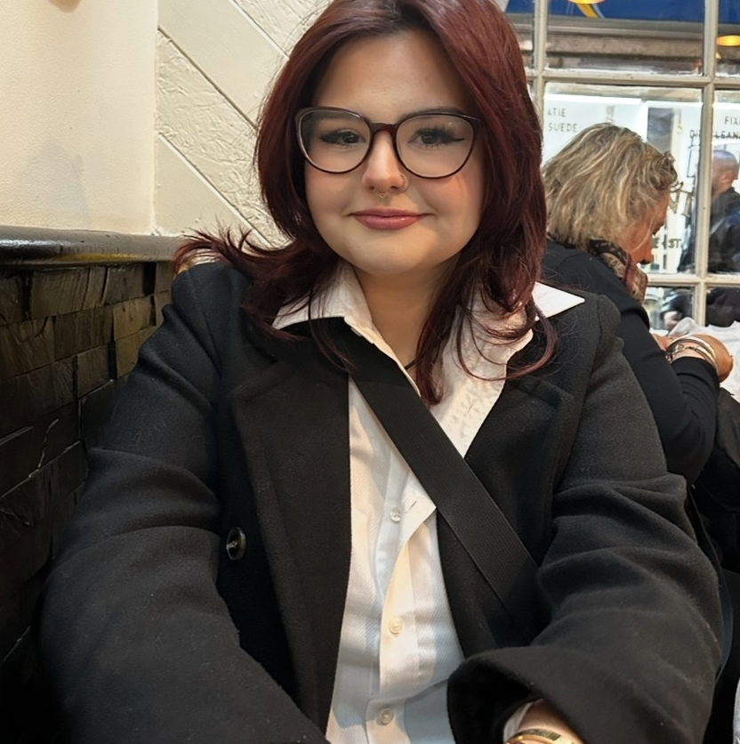 Leah is wearing a black coat and white shirt. She has shoulder length reddish hair and glasses