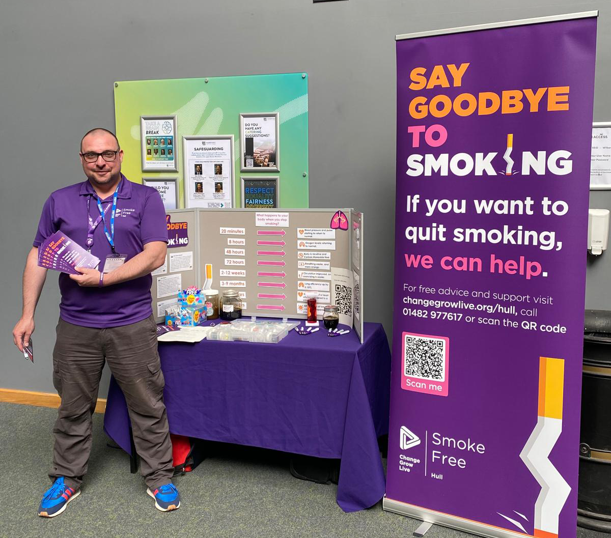 Sam is stood next to a table and banner with various leaflets and materials around stopping smoking