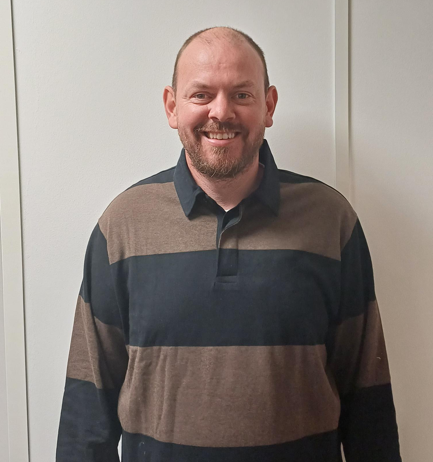 Simon is wearing a grey and blue long sleeved collared top. He has a moustache