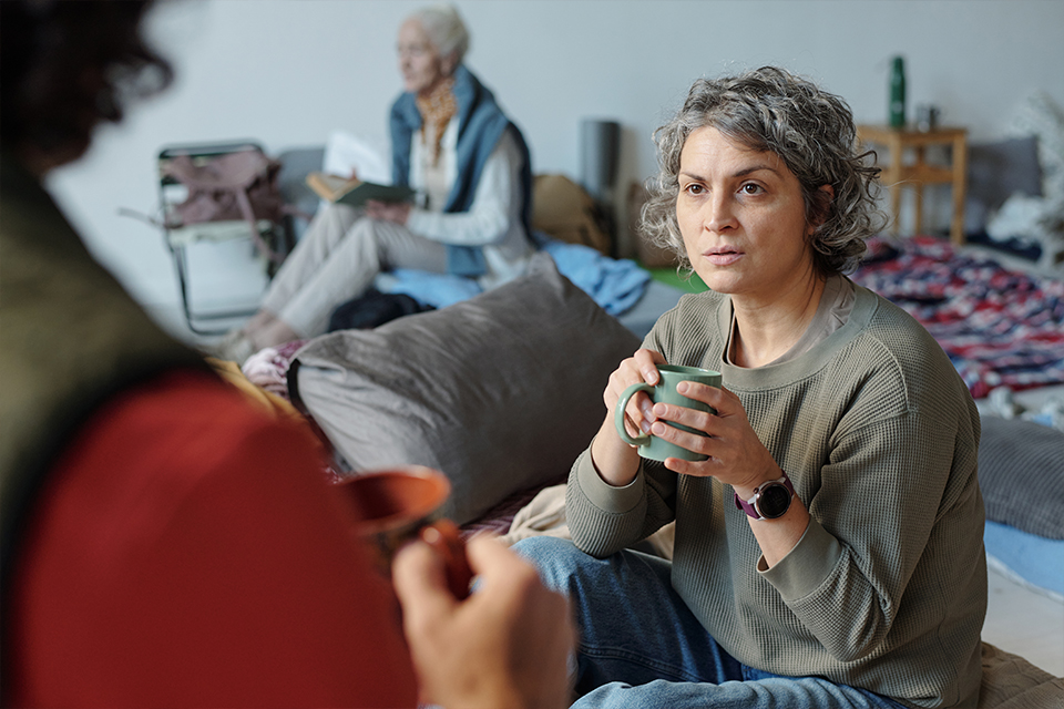 A stock photo of woman in homeless accommodation holding a cup of coffee and engaging in conversation with a friend