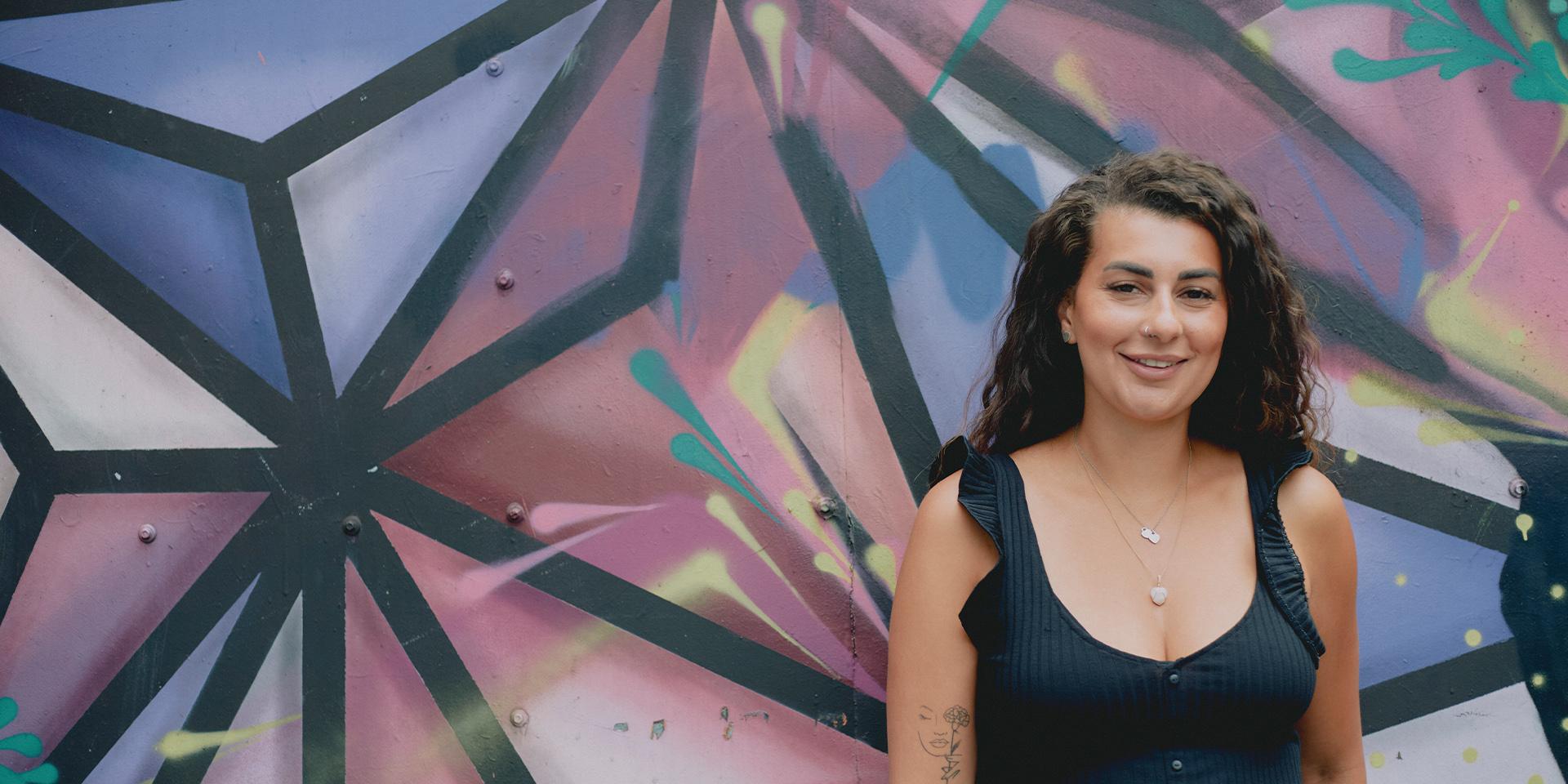 A person smiling at the camera with a mural in the background