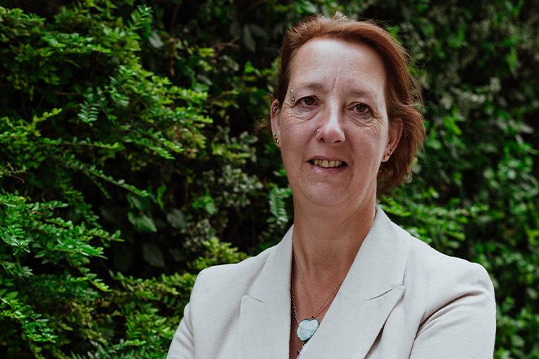 Debbie has short brown hair and is standing in front of green trees