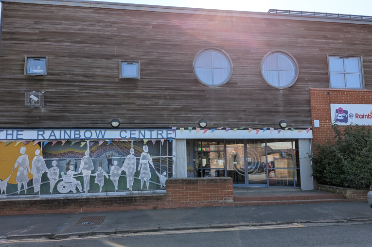 The outside of a building. On the ground floor is a wall length window with paper cut outs of people. The top half of the building has wooden panelling